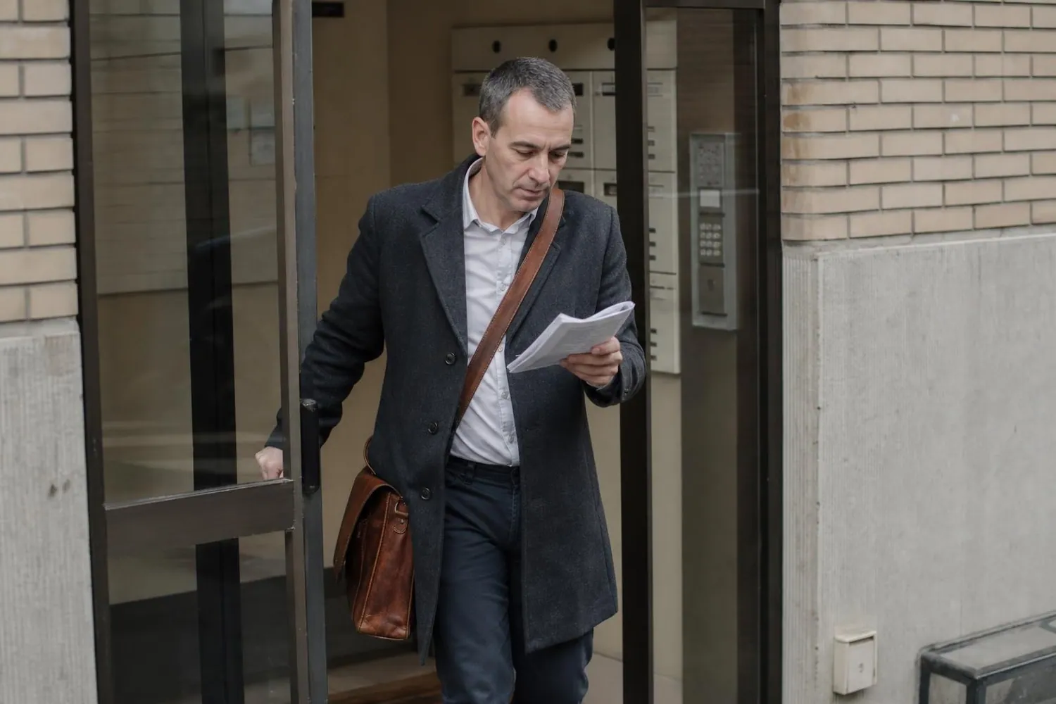 Clerc significateur en manteau sombre sortant du hall d une residence de logement social au petit matin, sacoche de cuir a l epaule